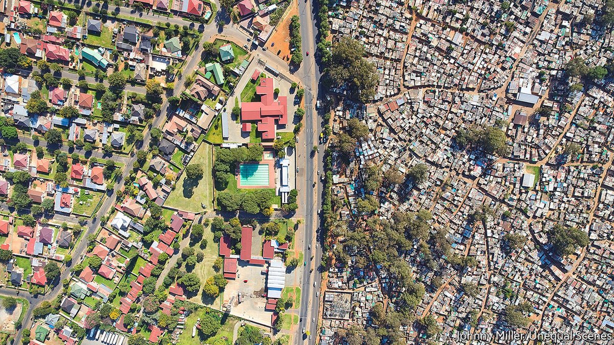 Aerial drone photograph divided by a road: left side shows an affluent suburban neighborhood with red-roofed houses, green lawns, a swimming pool, and a school complex; right side depicts a dense informal settlement (shack township) with corrugated metal structures, dirt paths, and limited vegetation, illustrating stark urban inequality.