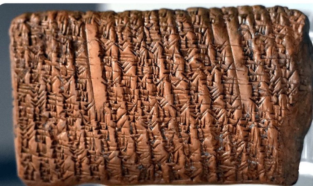 A rectangular, brownish clay tablet inscribed with dense wedge-shaped cuneiform markings, displayed on a dark surface with subtle lighting highlighting the textures and cracks of the artifact.