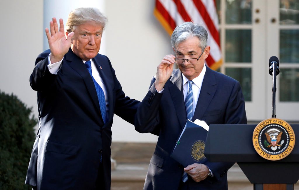 A formal outdoor photograph showing two men in suits standing at a White House podium: one (blond-haired, waving) is former President Donald Trump, and the other (gray-haired, adjusting eyeglasses while holding a blue folder) is Federal Reserve Chair Jerome Powell. An American flag and White House columns are in the background, with a presidential seal on the podium.