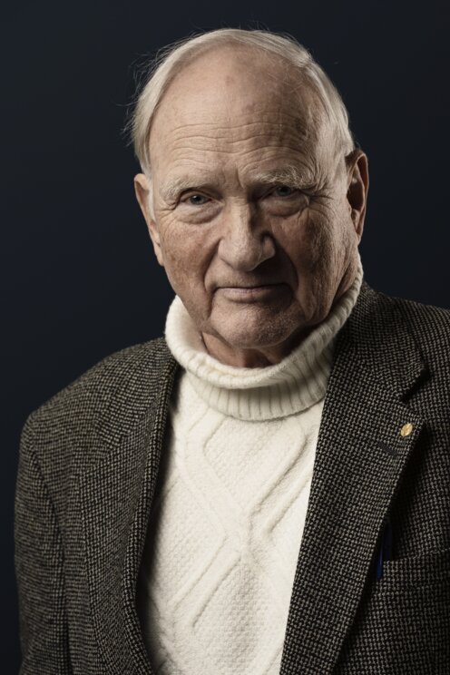 A formal portrait photograph of an elderly Caucasian man with short white hair, wearing a white cable-knit turtleneck sweater under a brown tweed jacket with a lapel pin, set against a dark blue background; he has a serious expression and is looking directly at the camera.